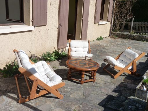 Un coin après repas. Trois chaises longues en bois et coussins autour d'une table en bois sur un patio ensoleillé.